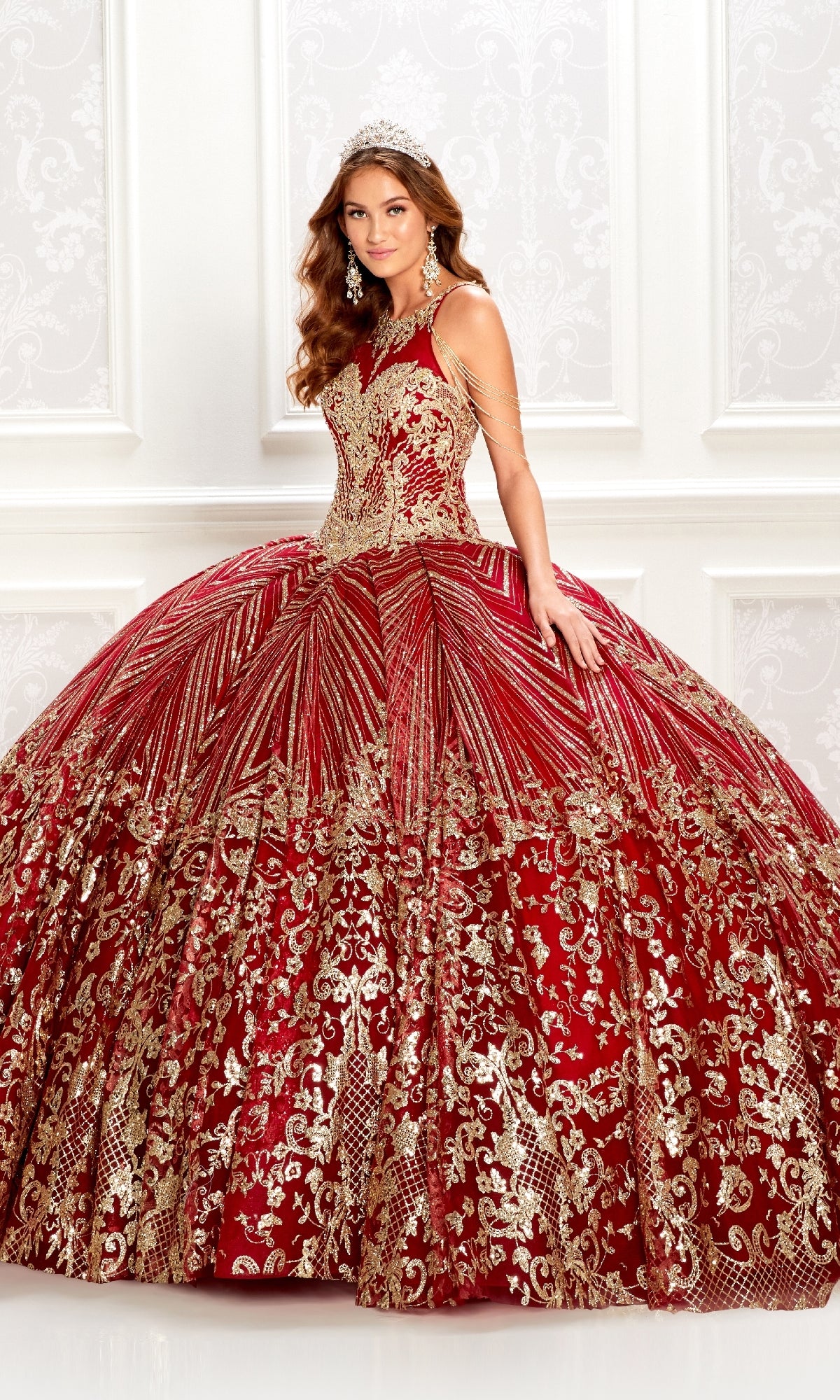 A young woman in the Princesa PR22034 wine red quinceanera dress with gold embellishments and a jeweled tiara stands before a white patterned wall.